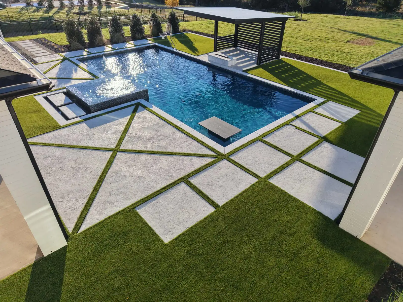Elevated view of a geometric pool with a raised spa, floating table, and turf-and-paver pattern beside a slatted pavilion in a Celina, TX backyard built by Prestige Pool & Patio