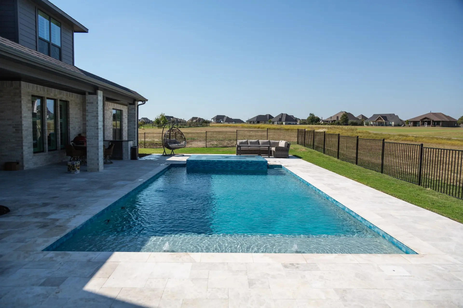 Rectangular pool with a raised tile spa, travertine decking, and a covered patio with seating in a Celina, TX backyard built by Prestige Pool & Patio