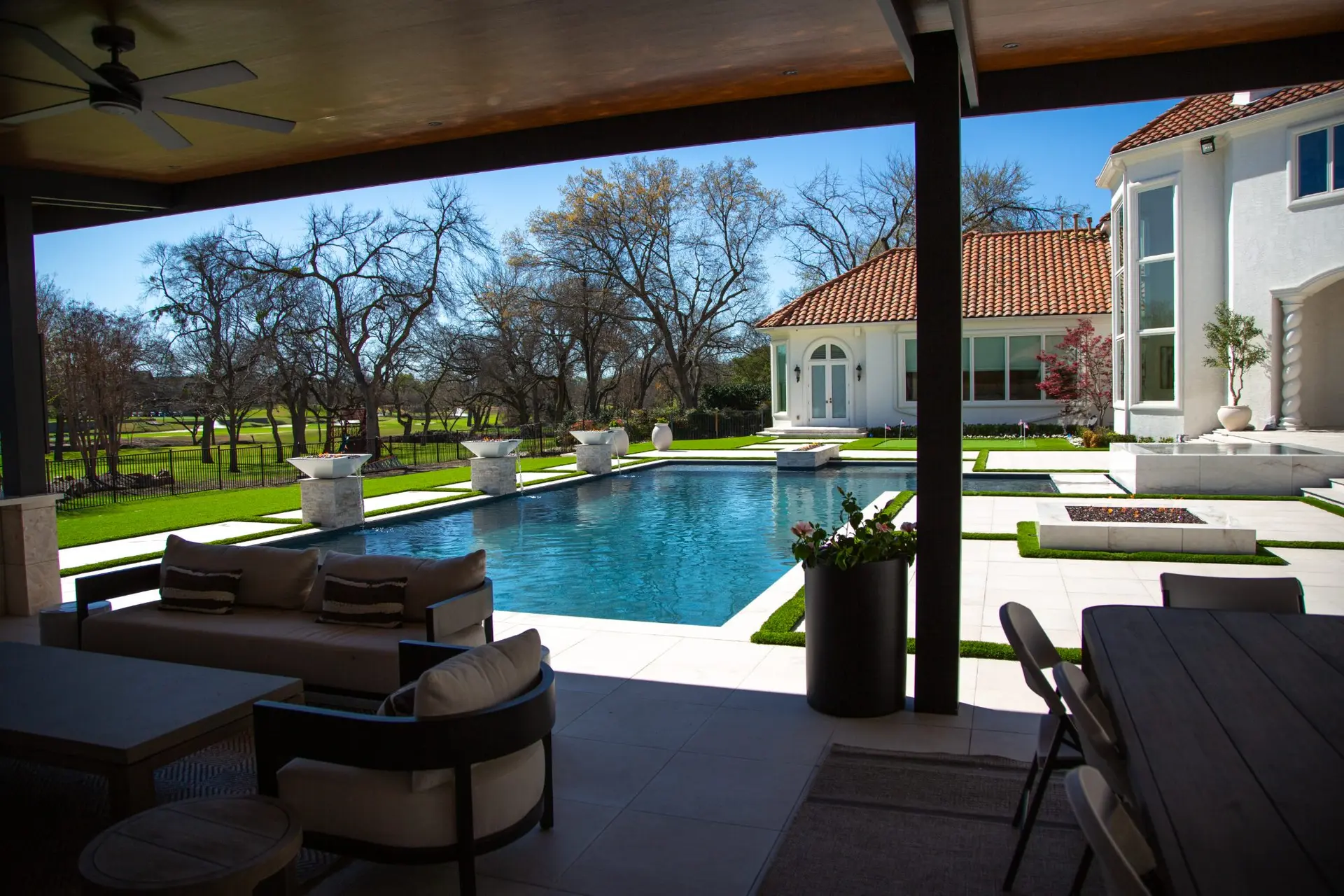 View from a covered patio of a rectangular pool with fire bowls and turf strips, looking out to mature trees in a North Texas backyard built by Prestige Pool & Patio