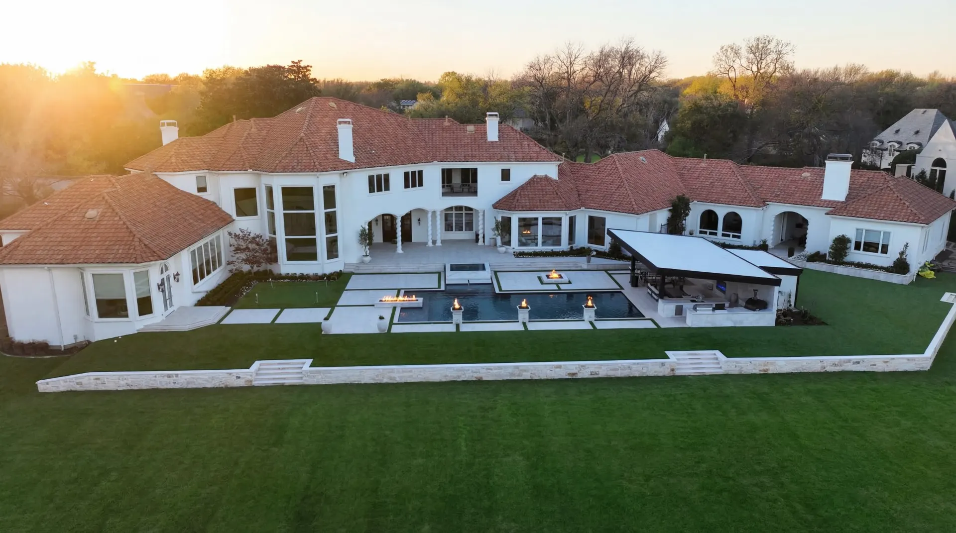 Aerial view at sunset of a geometric pool with a raised spa, fire bowls, and a covered outdoor kitchen behind a large Mediterranean-style home in a Plano, TX backyard built by Prestige Pool & Patio