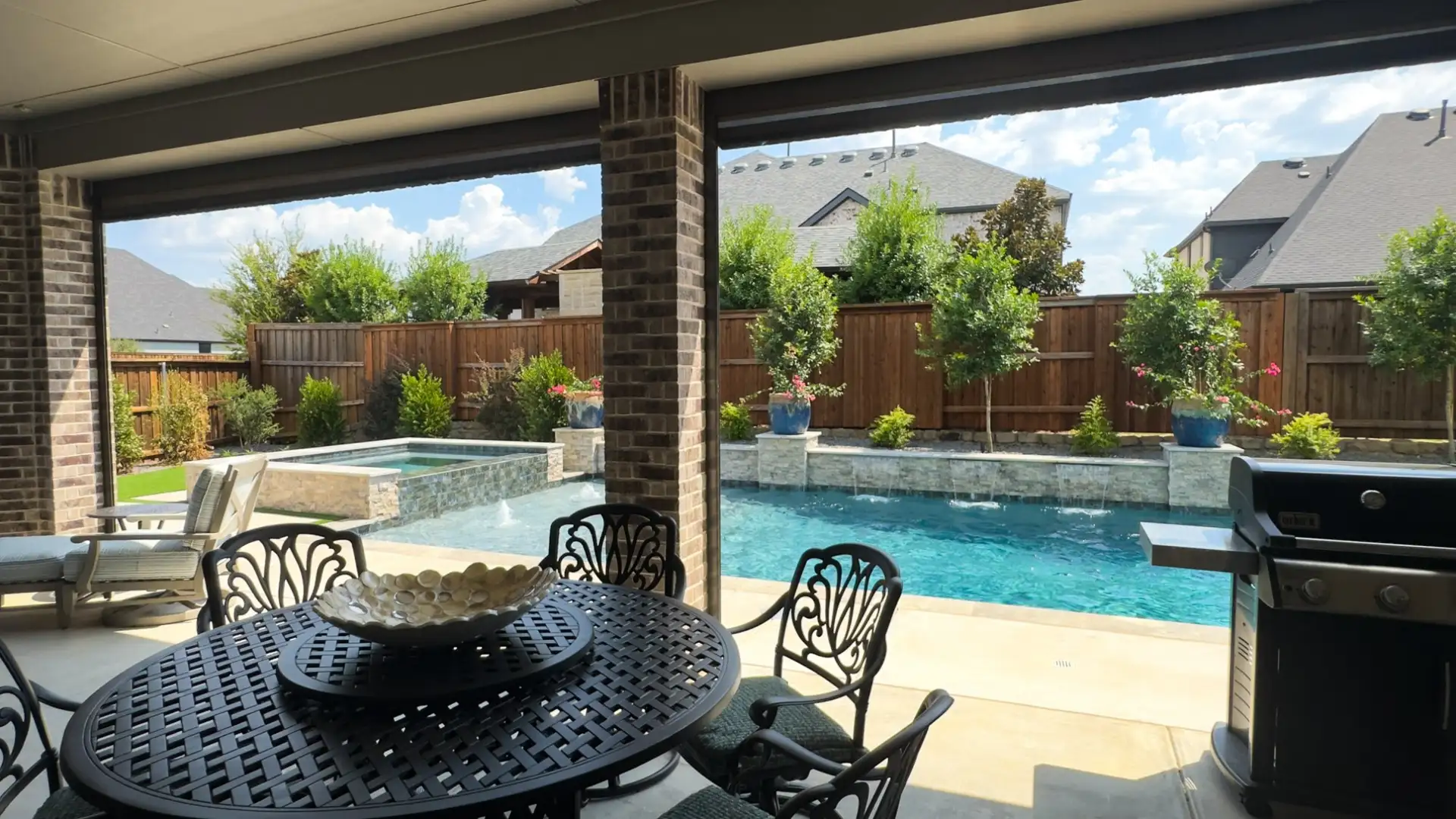 Covered patio dining area looking out to a geometric pool with a raised spa and sheer-descent water features in a Prosper, TX backyard built by Prestige Pool & Patio
