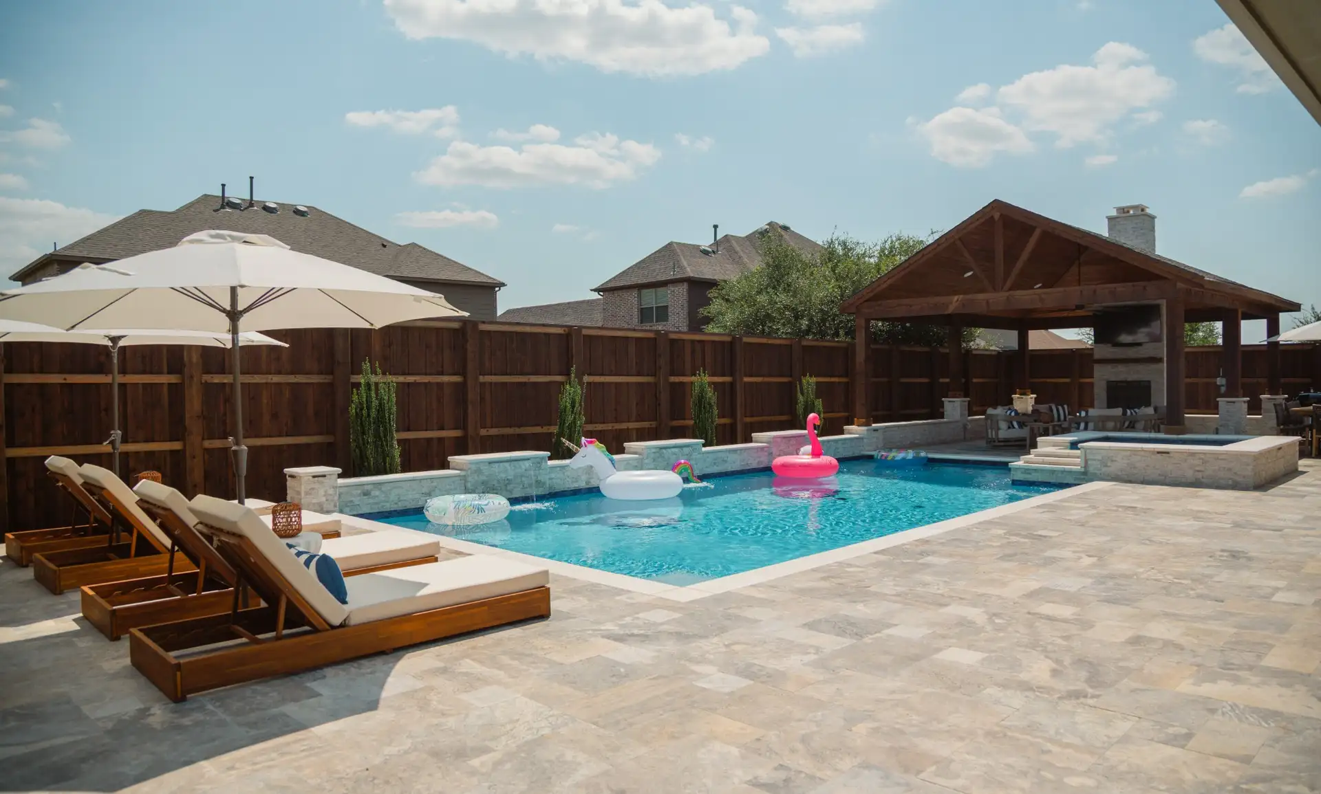 Geometric pool with a travertine deck, water features, raised spa, and covered pavilion in a Prosper, TX backyard built by Prestige Pool & Patio