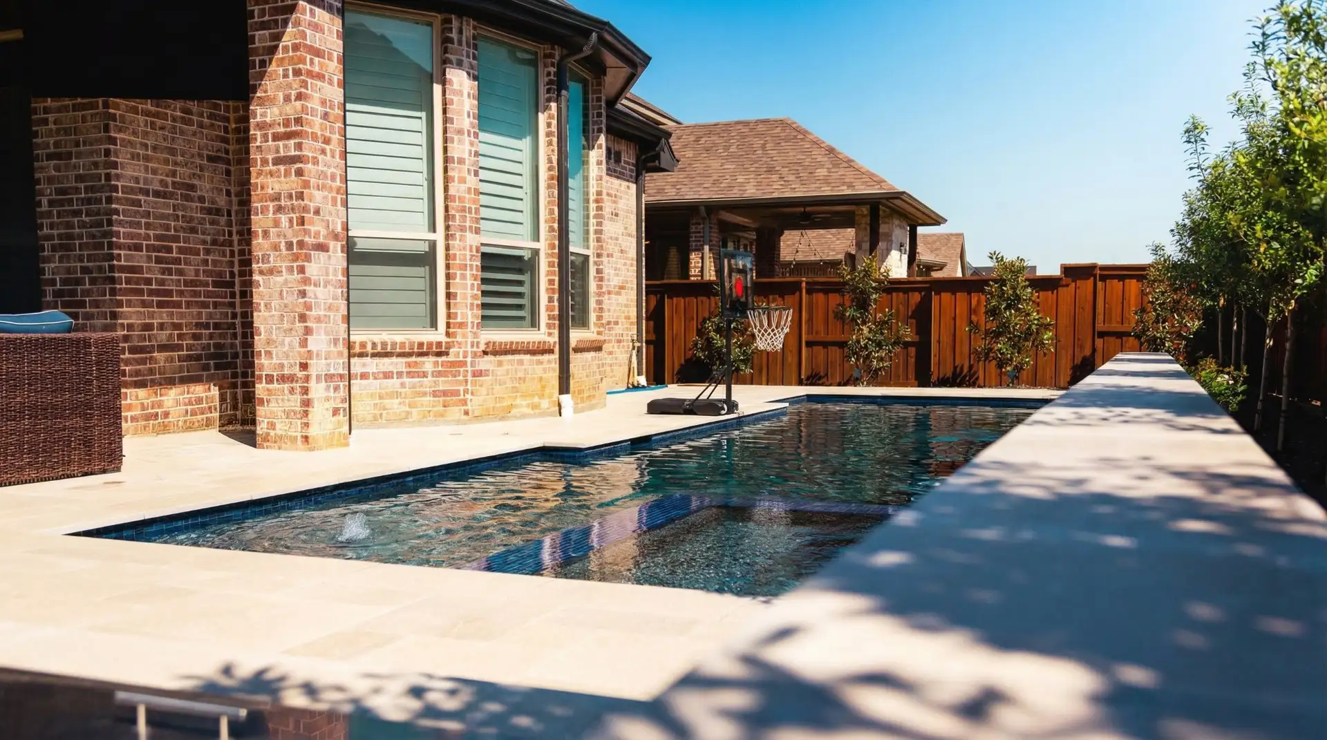 Long geometric pool with a tanning ledge and bubbler beside a brick home in a Prosper, TX backyard built by Prestige Pool & Patio